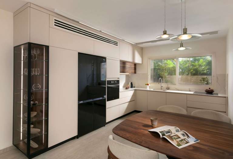 This modern kitchen is part of an open-plan living space, where the kitchen is designed as an integrated element within the overall layout. The design combines Sahara finish cabinetry, thin porcelain surfaces, black metal profiles and American walnut veneer, creating a precise and balanced material composition.